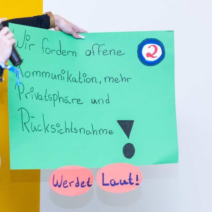 Junger Mensch hält auf der Bühne ein Plakat mit der Aufschrift: "Wir fordern offene Kommunikation, mehr Privatsphäre und Rücksichtnahme! Werdet laut!" (vergrößerte Bildansicht wird geöffnet)
