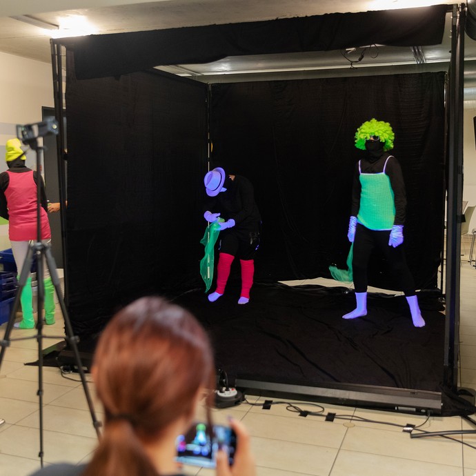 Das Bild zeigt Personen in einer schwarzen Bühne, die neonfarbene Kleidung tragen und von Schwarzlicht beleuchtet werden (vergrößerte Bildansicht wird geöffnet)