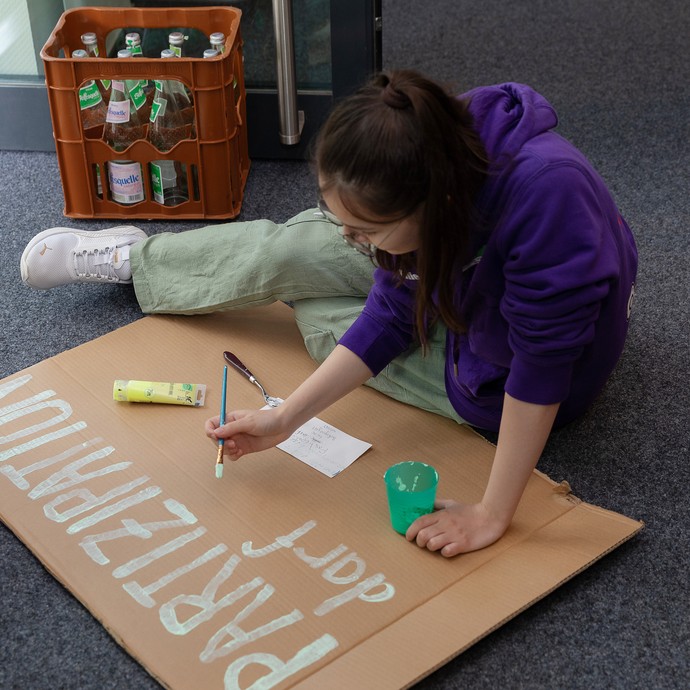 Das Bild zeigt eine Person, die auf dem Boden sitzt und ein Papp-Plakat beschreibt. Zu lesen ist "Partizipation darf" (vergrößerte Bildansicht wird geöffnet)