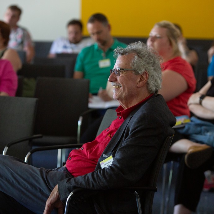 Das Bild zeigt Prof. Dr. Klaus Wolf, der lachend zwischen Fachkräften sitzt (vergrößerte Bildansicht wird geöffnet)