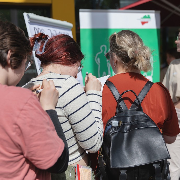 Das Bild zeigt 3 Personen, die in einer Reihe stehen und den Rücken der vorderen Person als Schreibunterlage nutzen (vergrößerte Bildansicht wird geöffnet)