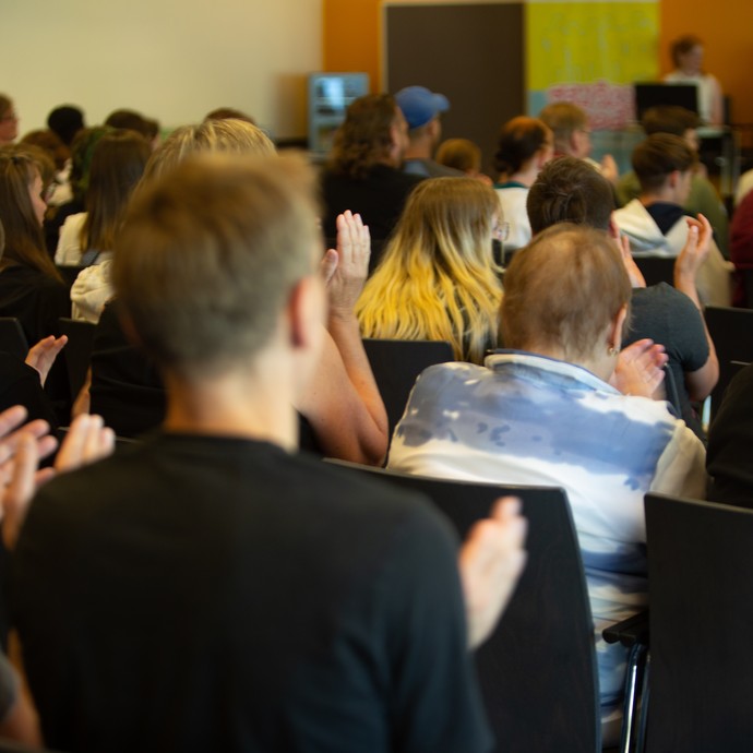 Das Bild zeigt das Publikum von hinten, das auf die Bühne schaut (vergrößerte Bildansicht wird geöffnet)