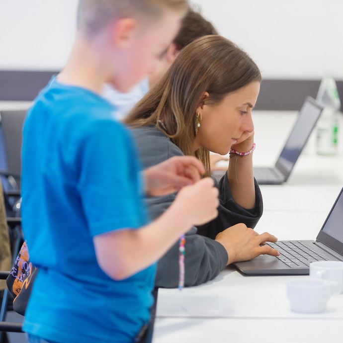 Das Bild zeigt ein Kind und eine erwachsene Person, die vor einem Laptop sitzen (vergrößerte Bildansicht wird geöffnet)