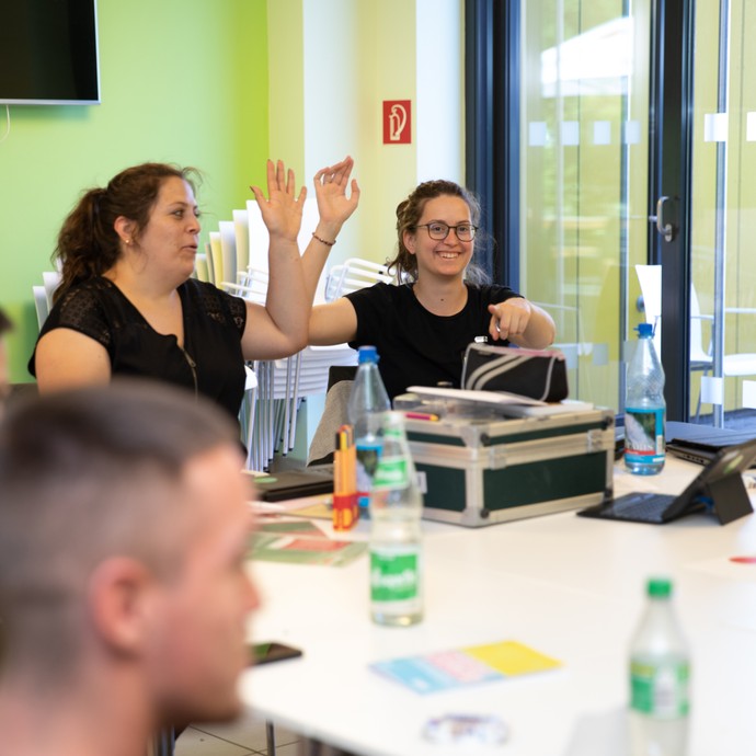 Das Bild zeigt zwei Workshop-Leiterinnen, die lachend die Hand heben (vergrößerte Bildansicht wird geöffnet)