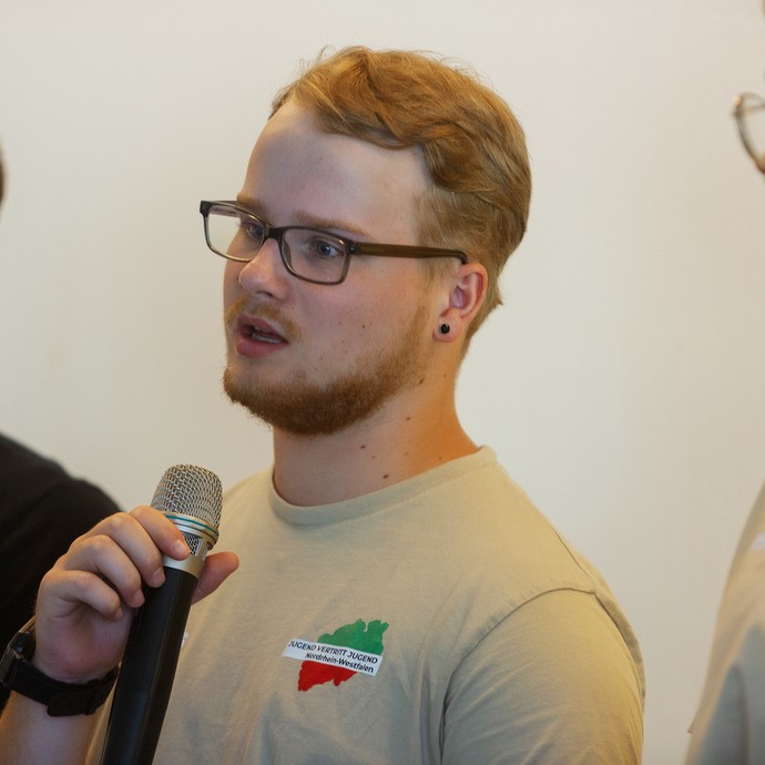 Das Bild zeigt ein JvJ-Mitglied mit Bart und Brille, das ein Mikro in der Hand hält (vergrößerte Bildansicht wird geöffnet)