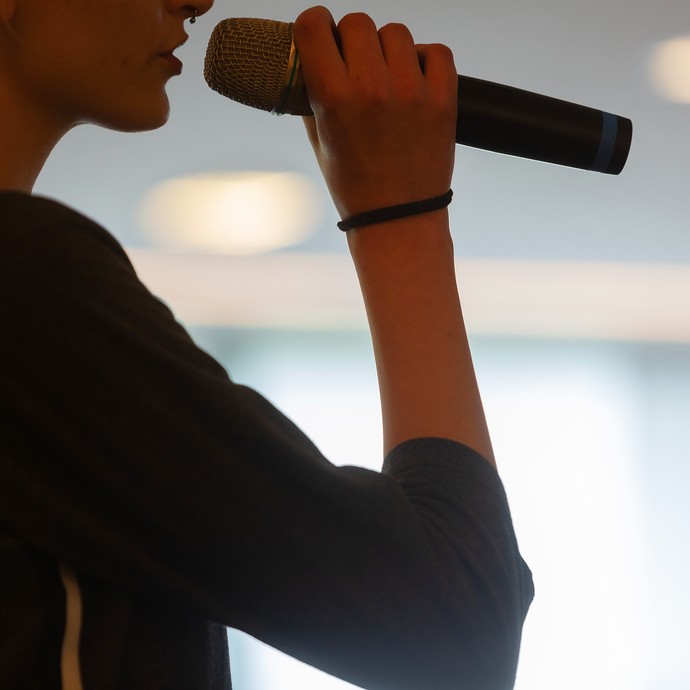 Das Bild zeigt eine Silhouette einer Person mit einem Mikrofon in der Hand (vergrößerte Bildansicht wird geöffnet)