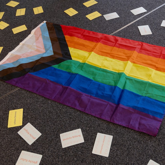 Auf dem Boden liegen eine bunte LQBTQIA+-Flagge und verschiedene Karten. (vergrößerte Bildansicht wird geöffnet)